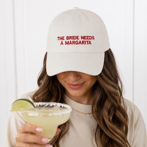 May include: An off-white baseball cap featuring the embroidered red text THE BRIDE NEEDS A MARGARITA on the front panel. The cap has a structured crown, a curved brim, and a matching fabric strap with an adjustable metal buckle at the back.