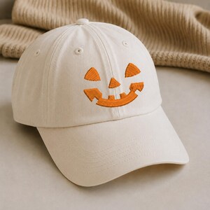 Peut inclure: Une casquette de baseball blanc cassé avec un visage de citrouille d'Halloween orange brodé sur le panneau avant. La casquette présente une calotte structurée, une visière incurvée et des œillets cousus, avec une construction classique à six panneaux.