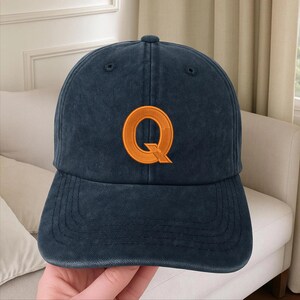 Op de afbeelding: Een donker marineblauwe baseballpet met een verweerde, verwassen textuur. Het voorpaneel is voorzien van een grote, geborduurde hoofdletter Q in fel oranje draad met een opvallende, gestileerde diagonale streep rechtsonder.