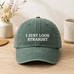 Può includere: Un cappellino da baseball verde bosco con una texture slavata e vissuta. Il pannello frontale presenta il testo ricamato in bianco "I JUST LOOK STRAIGHT" in un carattere sans-serif pulito. Il cappello ha una corona strutturata e una visiera.