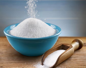 May include: A blue ceramic bowl filled with white granulated sugar, with more sugar being poured into it. A wooden scoop filled with sugar sits beside the bowl on a wooden surface.
