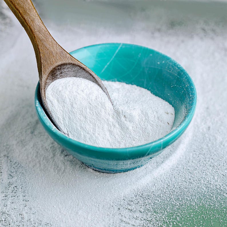 May include: A close-up shot of a teal bowl filled with white powder, with a wooden spoon partially submerged. The powder spills out onto the surface. The bowl is round and has a slightly textured surface.