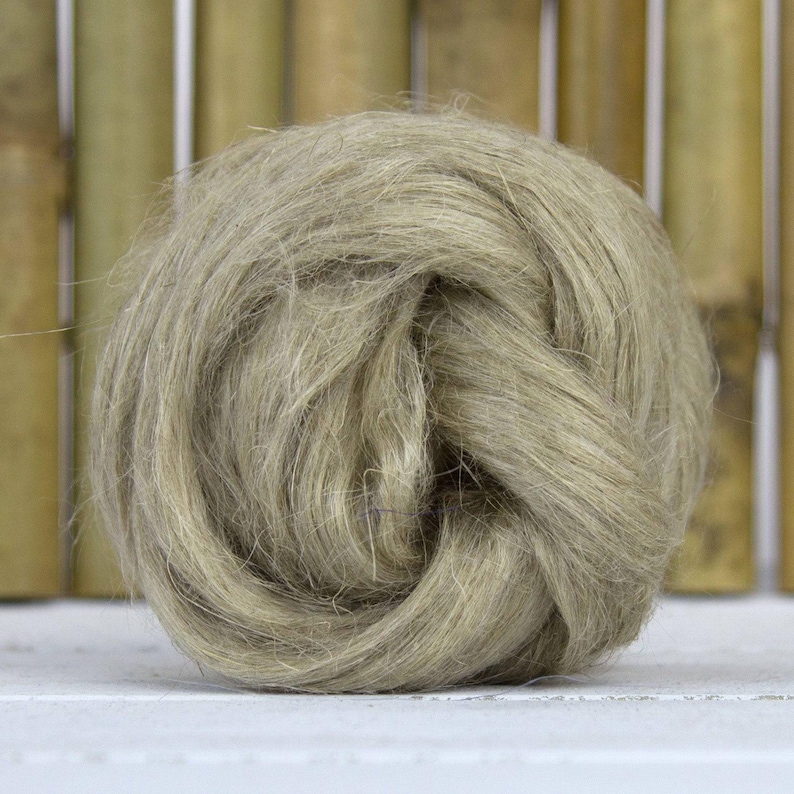 Puede incluir: Una bola de fibra de lino de color natural, bien enrollada y texturizada. Las fibras son de un tono apagado y terroso, con una forma ligeramente irregular. El fondo presenta ca&ntilde;as de bamb&uacute; verticales, a&ntilde;adiendo un elemento natural. Materias primas para la artesan&iacute;a.