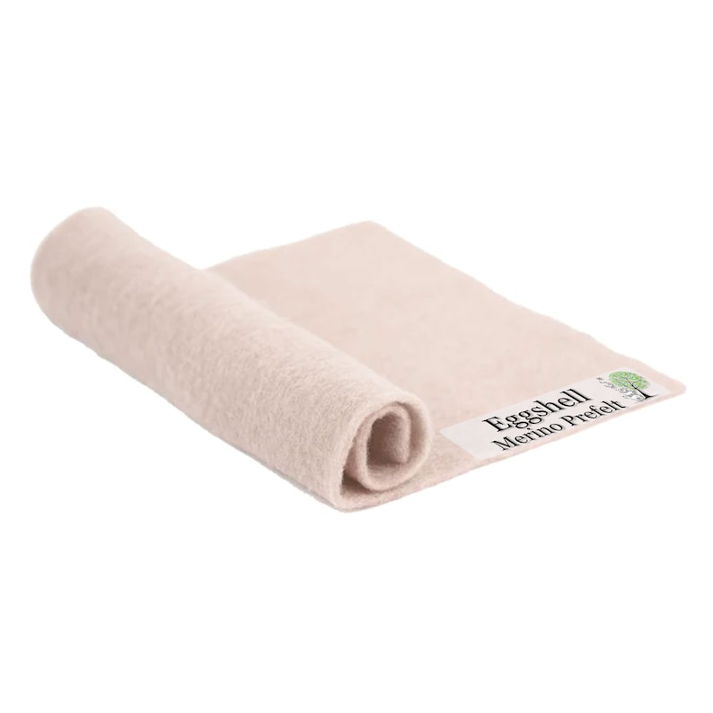 May include: A rolled-up, off-white merino wool prefelt. The fabric is a soft, neutral color, with a slightly textured surface. The label reads "Eggshell Merino Prefelt" with a tree logo.