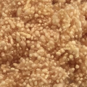 May include: Close-up of a pile of beige, fluffy material. The texture is dense with small, round, light-colored nodules interspersed throughout. The overall appearance suggests a soft, natural fiber, possibly wool or a similar textile.