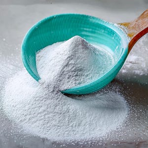 May include: A turquoise ceramic bowl with a wooden handle is filled with a mound of white powder, some of which has spilled out onto the surface. The bowl is tilted, suggesting the powder is being poured or measured. The image is well-lit, highlighting the texture of the powder.