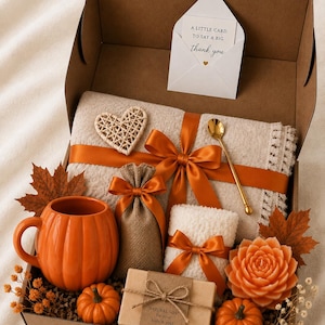 Könnte beinhalten: Eine Geschenkbox mit einer beigen Strickdecke mit einem rostorangen Band, einer kürbisförmigen orangefarbenen Tasse, einem kleinen Jutebeutel, einem weißen flauschigen Gegenstand, einem Stück Seife mit der Aufschrift Natural Soap, zwei kleinen Kürbissen, einer blumenförmigen Kerze und einer Karte.