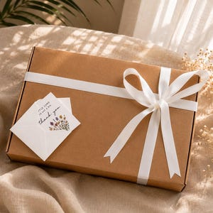 Puede incluir: Una caja de regalo rectangular de cart&oacute;n marr&oacute;n envuelta con una cinta de sat&eacute;n blanca atada en un lazo. Un peque&ntilde;o sobre blanco est&aacute; metido bajo la cinta, con una ilustraci&oacute;n de flores silvestres y el texto, "A little card to say a big thank you."
