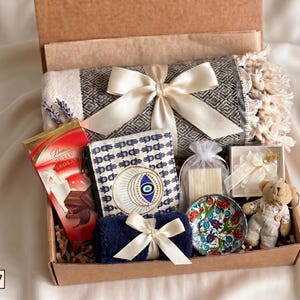 Puede incluir: Una caja de regalo que contiene una manta gris estampada con una cinta color crema, una barra de chocolate roja, una bolsa con estampado de hamsa y ojo, una toalla azul peque&ntilde;a con cinta crema, un cuenco de cer&aacute;mica floral, una vela y un osito.