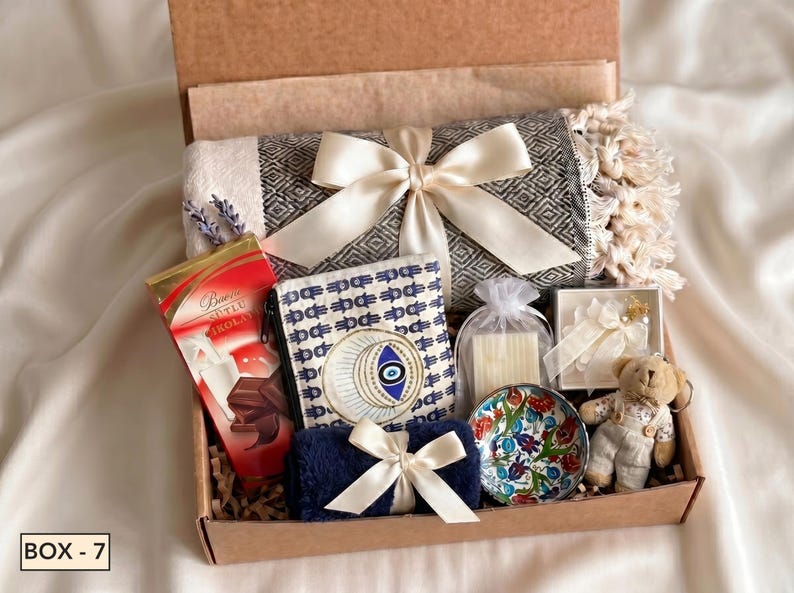 Puede incluir: Una caja de regalo que contiene una manta gris estampada con una cinta color crema, una barra de chocolate roja, una bolsa con estampado de hamsa y ojo, una toalla azul peque&ntilde;a con cinta crema, un cuenco de cer&aacute;mica floral, una vela y un osito.