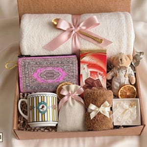Puede incluir: Una caja de regalo que contiene una manta polar blanca con un lazo rosa, una cuchara dorada, un estuche con estampado morado, una caja de chocolate, un osito de peluche, una taza a rayas con un p&aacute;jaro, una bolsa beige y fruta deshidratada.
