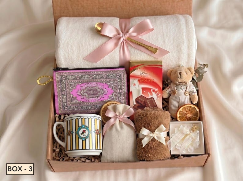 Puede incluir: Una caja de regalo que contiene una manta polar blanca con un lazo rosa, una cuchara dorada, un estuche con estampado morado, una caja de chocolate, un osito de peluche, una taza a rayas con un p&aacute;jaro, una bolsa beige y fruta deshidratada.