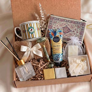 May include: A gift box containing a striped mug with a bird motif, a patterned zippered pouch, a tube of Blue Moon hand and body cream, a green jade facial roller, a glass reed diffuser, a small white organza bag, and a white floral hair accessory.