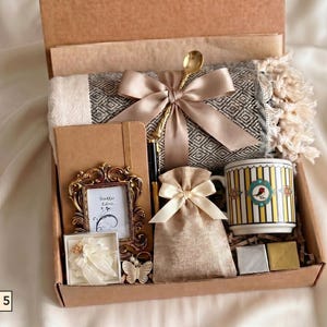 Puede incluir: Una caja de regalo que contiene una manta con patr&oacute;n gris y blanco con una cinta beige, un cuaderno marr&oacute;n, una cuchara dorada, un peque&ntilde;o marco dorado ornamentado, una bolsa beige, una taza con rayas amarillas y un p&aacute;jaro, y dos cuadrados peque&ntilde;os.