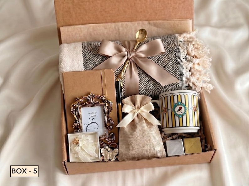 Puede incluir: Una caja de regalo que contiene una manta con patr&oacute;n gris y blanco con una cinta beige, un cuaderno marr&oacute;n, una cuchara dorada, un peque&ntilde;o marco dorado ornamentado, una bolsa beige, una taza con rayas amarillas y un p&aacute;jaro, y dos cuadrados peque&ntilde;os.