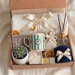 Puede incluir: Una caja de regalo que contiene una manta blanca con un lazo crema, una cuchara dorada, una taza con rayas azules y un motivo de p&aacute;jaro, una suculenta en maceta blanca, un difusor de varillas, una bolsita, una toalla azul marino y fruta seca.