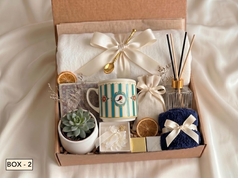 Puede incluir: Una caja de regalo que contiene una manta blanca con un lazo crema, una cuchara dorada, una taza con rayas azules y un motivo de p&aacute;jaro, una suculenta en maceta blanca, un difusor de varillas, una bolsita, una toalla azul marino y fruta seca.