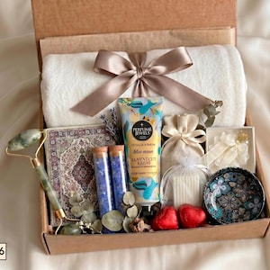 Puede incluir: Una caja de regalo que contiene una manta de felpa blanca con una cinta color topo, un rodillo facial de jade verde, un cuaderno estampado, dos tubos de vidrio con sales de ba&ntilde;o azules, crema Perfume Jewels Blue Moon, dos jabones de coraz&oacute;n rojos.