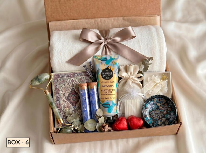 Puede incluir: Una caja de regalo que contiene una manta de felpa blanca con una cinta color topo, un rodillo facial de jade verde, un cuaderno estampado, dos tubos de vidrio con sales de ba&ntilde;o azules, crema Perfume Jewels Blue Moon, dos jabones de coraz&oacute;n rojos.