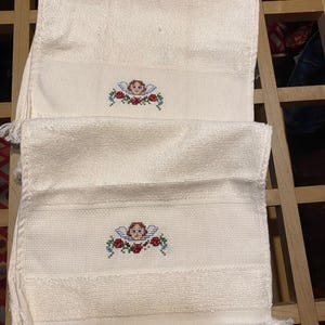 May include: Two off-white, fringed hand towels. Each towel features a cross-stitched design of an angel with blue wings and a wreath of red roses and blue flowers. The towels are folded and stacked.