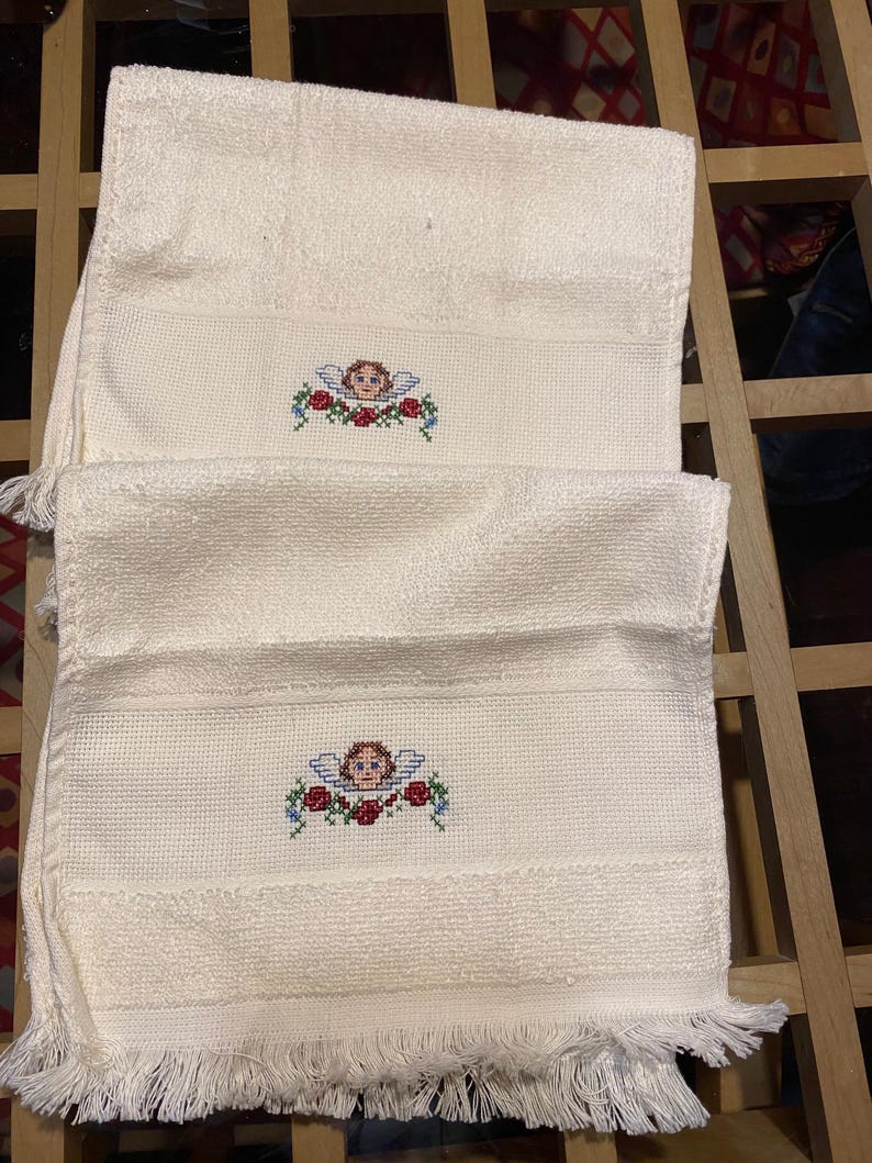 May include: Two off-white, fringed hand towels. Each towel features a cross-stitched design of an angel with blue wings and a wreath of red roses and blue flowers. The towels are folded and stacked.