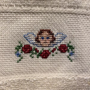 May include: Cream-colored towel with a cross-stitched design featuring an angel with brown hair, blue eyes, and blue wings. The angel is surrounded by red roses and green foliage. The towel has a textured surface.
