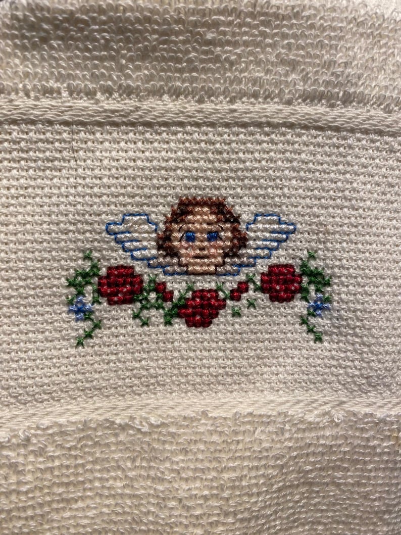 May include: Cream-colored towel with a cross-stitched design featuring an angel with brown hair, blue eyes, and blue wings. The angel is surrounded by red roses and green foliage. The towel has a textured surface.