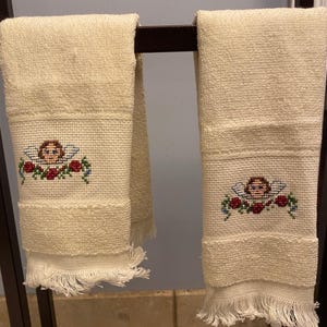 May include: Two cream-colored hand towels with a cross-stitched design of an angel with wings and a floral garland. The towels have a fringed edge and are hanging on a dark metal rack. The towels are made of a textured fabric.