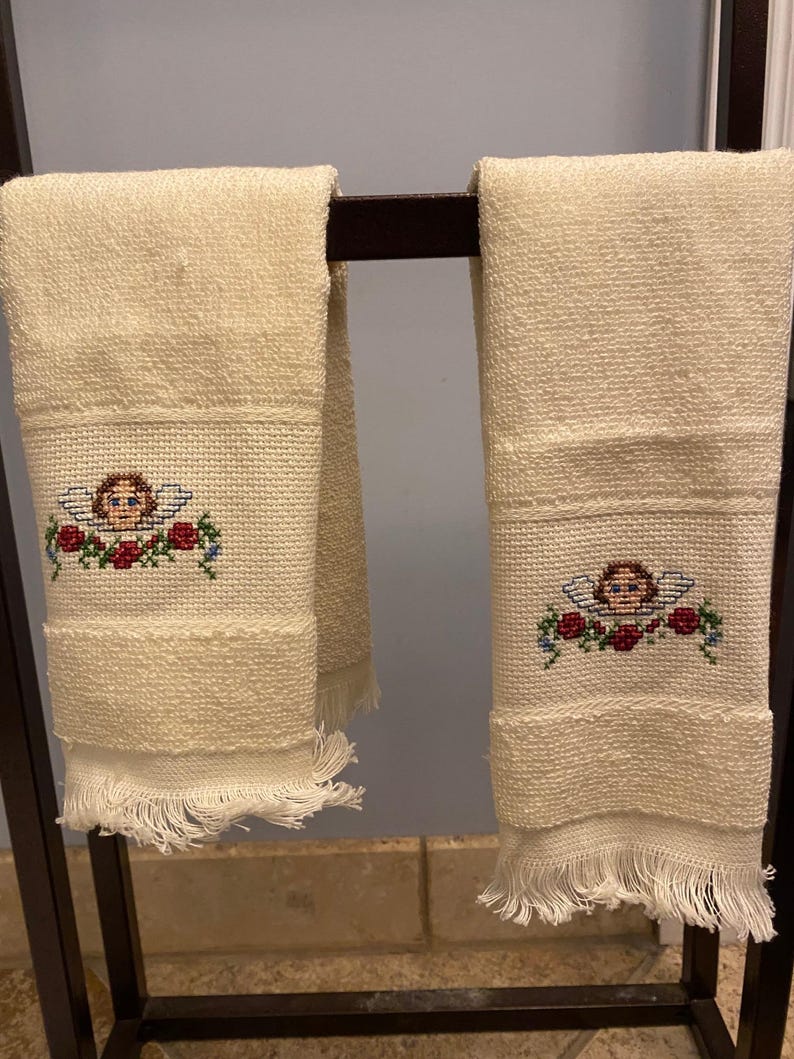 May include: Two cream-colored hand towels with a cross-stitched design of an angel with wings and a floral garland. The towels have a fringed edge and are hanging on a dark metal rack. The towels are made of a textured fabric.