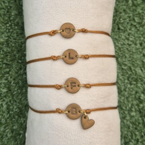 Op de afbeelding: Vier armbanden met dunne bruine koordjes en ronde houten bedels. De bedels zijn gegraveerd met een hartje, de letter L, de letter F en de letter B. De onderste armband bevat ook een kleine houten bedel in de vorm van een hartje.