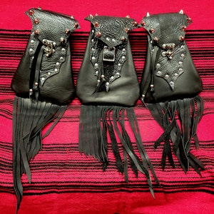 Pode incluir: Três bolsas de couro preto com franjas, tachas de metal e detalhes decorativos. Cada bolsa tem um fecho de aba e uma longa franja. As bolsas são exibidas em um tecido listrado vermelho e preto.