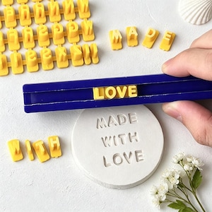 May include: A set of yellow plastic alphabet letter stamps and a blue plastic holder bar. The stamps are used to press the words MADE WITH LOVE into a circular white clay disc. The letters are arranged in the holder to spell the word LOVE.