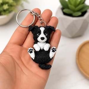 May include: A keychain featuring a sculpted black and white dog with a white blaze on its face, white chest, and white paws with black paw pads. The dog is in a seated position with its front paws raised, attached to a circular silver metal ring.