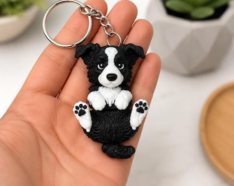 Könnte beinhalten: Ein schwarz-weißer Border Collie Schlüsselanhänger mit einer skulptierten Figur, die eine weiße Brust, Pfoten und Gesichtsmarkierungen aufweist und an einem silberfarbenen Metallring mit einer kurzen Kette befestigt ist. Der Hund liegt auf dem Rücken.