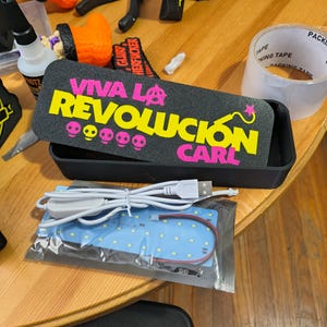 May include: A black rectangular display box featuring a sign with the text VIVA LA REVOLUCION CARL in yellow and pink, including an anarchy symbol and skull icons. Below the box is a blue LED light strip with a white USB power cable.