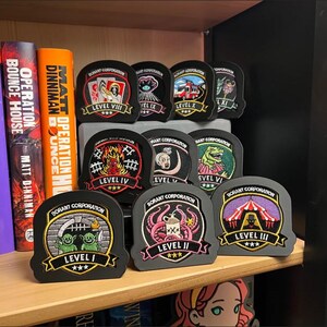 May include: A collection of Borant Corporation embroidered patches displayed on black stands. Each patch features a unique graphic design, a level number from I to XI, and three stars. The patches are arranged in rows on a wooden shelf next to books.