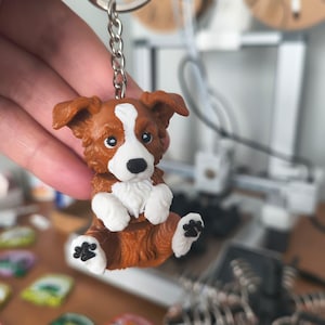 May include: A brown and white dog figurine keychain featuring a puppy with floppy ears, a white blaze on its face, and white paws with black paw pad details. The dog is attached to a silver-colored metal split ring by a short metal chain.