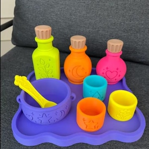 May include: A set of silicone play items including a purple tray, a purple cauldron with a yellow spoon, three bottles with tan stoppers in lime green, orange, and pink, and three small cups in blue, orange, and yellow with embossed icons.