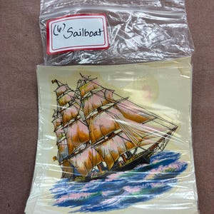 May include: A stack of square paper decals featuring a multi-masted sailboat with orange-tinted sails on blue and white waves, enclosed in a clear plastic zip-top bag with a white label reading (6) Sailboat in black ink on the front.