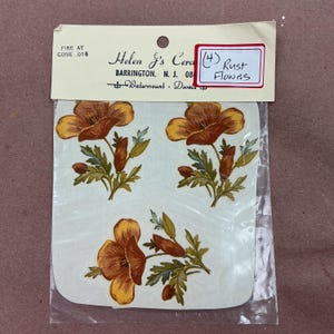 May include: A package of three water-mount ceramic decals featuring rust-colored flowers with green leaves. The header card reads Helen J's Ceramics, Barrington, N.J., Fire at Cone .016, and includes a handwritten label stating (4) Rust Flowers.