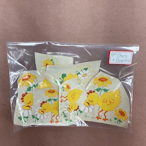 May include: A clear plastic bag containing multiple adhesive decals featuring yellow chicks and white and yellow flowers. A white label on the bag has the handwritten text "(6) Chics + flowers" in black ink. A ruler is visible at the bottom edge.