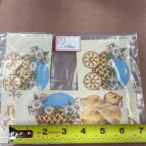 May include: Three paper cutouts featuring Easter themes, including a yellow chick and woven baskets with daisies and blue eggs. A yellow Stanley measuring tape is placed along the bottom, showing a width of approximately 6.5 inches.