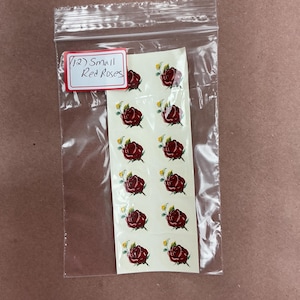 May include: A clear plastic resealable bag containing a white rectangular sheet with twelve small red rose stickers. Each rose features dark red petals and small green leaves. A red-bordered white label at the top reads (12) Small Red Roses.
