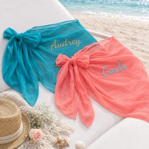 May include: Two sheer sarongs, one teal and one coral, each tied in a knot. The teal sarong features the name Audrey embroidered in gold, while the coral sarong features the name Camila embroidered in blue. Both are made of lightweight fabric.