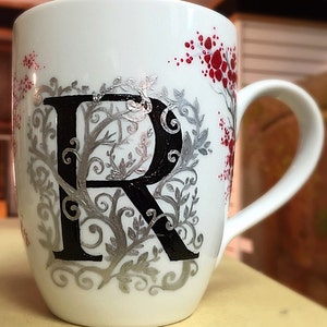 May include: White ceramic mug featuring a black letter "R" surrounded by silver and red floral designs.