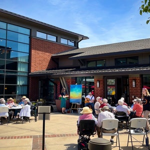 May include: A group of people gather outside a brick building with a large glass window. Some people are seated at tables, while others are listening to a musician playing a guitar. A painting is displayed on an easel in the background.