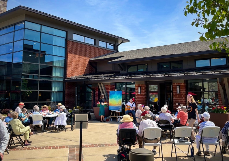 May include: A group of people gather outside a brick building with a large glass window. Some people are seated at tables, while others are listening to a musician playing a guitar. A painting is displayed on an easel in the background.