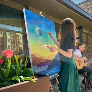 May include: A woman paints a landscape with a solar eclipse in the sky on a canvas easel. A man plays a guitar in the background.