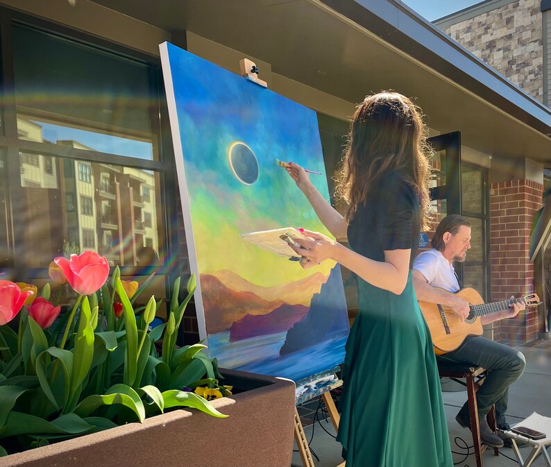 May include: A woman paints a landscape with a solar eclipse in the sky on a canvas easel. A man plays a guitar in the background.