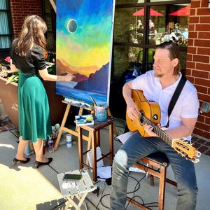 May include: A woman paints a landscape on canvas while a man plays an acoustic guitar. The painting depicts a sunset with a dark circle in the sky, possibly a solar eclipse. The scene is set outdoors, with brick walls and a window in the background.